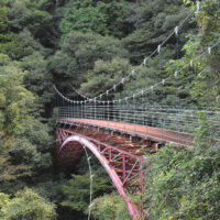 遠登志橋。赤いアーチ橋の部分は明治38年に完成。当時は橋の上流側に抗水路が併設されていた。平成5年にア―チ橋の上にワイヤーロープ吊橋が架設された。