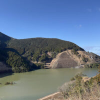 試験湛水中のあそ立野ダム：昭和28年(1953年)の熊本大水害を機に持ち上がったダム計画、洪水調節を目的とした流水型のダムで2023年完成し、2024年初め試験湛水が行われた。堤高87m、堤頂長197m、満水になると北向谷原始林の下部は水没する。
